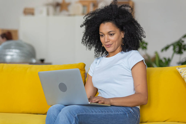 Portrait of a young African American woman typing on laptop keyboard. Brunette with curly hair sitting on yellow sofa in a bright home room. Close up. - Fotoğraf, Görsel