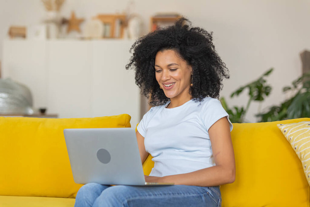 Portrait of a young African American woman talking on a video call on portable laptop. Brunette with curly hair sitting on yellow sofa in a bright home room. Close up. - Zdjęcie, obraz