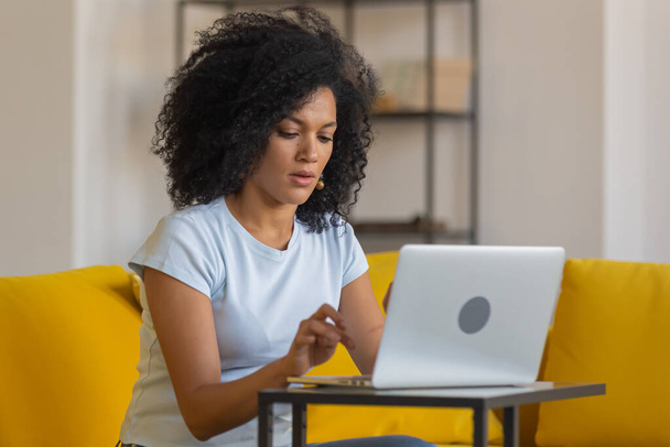 Portrait of a young African American woman typing on laptop keyboard. Brunette with curly hair sitting on yellow sofa in a bright home room. Close up. - Fotografie, Obrázek