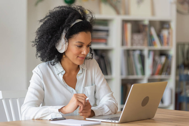 Portrait of African American woman talking on video conference call using laptop and headphones taking notes on notepad. Brunette sits at table in home office. Close up. - Foto, afbeelding