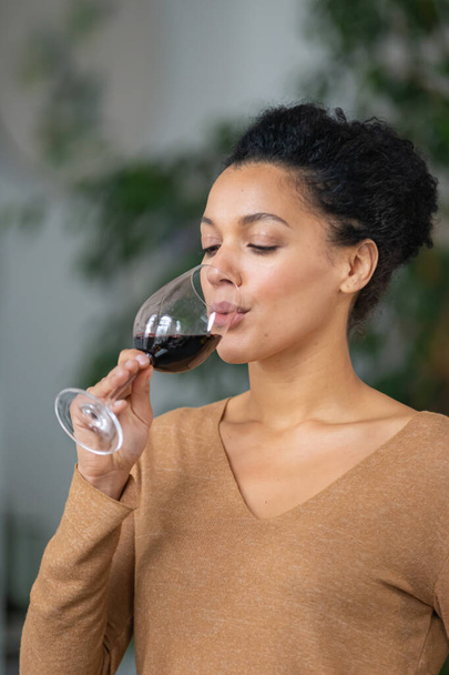 Portrait of a young African American woman drinking red wine from glass goblet. Cute mixed race female is enjoying taste of drink against blurred background of light room with green plants. Close up. - 写真・画像