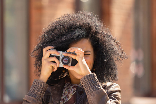Stylish young African American woman taking pictures with a retro camera. Brunette with curly hair in brown leather jacket poses against background of blurred brick building. Close up. - Photo, Image