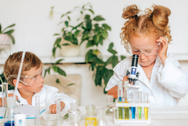 Boy and girl in white uniforms and protective glasses do chemical experiments in a home laboratory.Back to school concept.Young scientists.Natural sciences.Preschool and school education of children. - Φωτογραφία, εικόνα