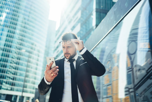 From below of confused male entrepreneur in stylish formal clothing taking of eyeglasses while browsing smartphone on sunny day at downtown  - 写真・画像