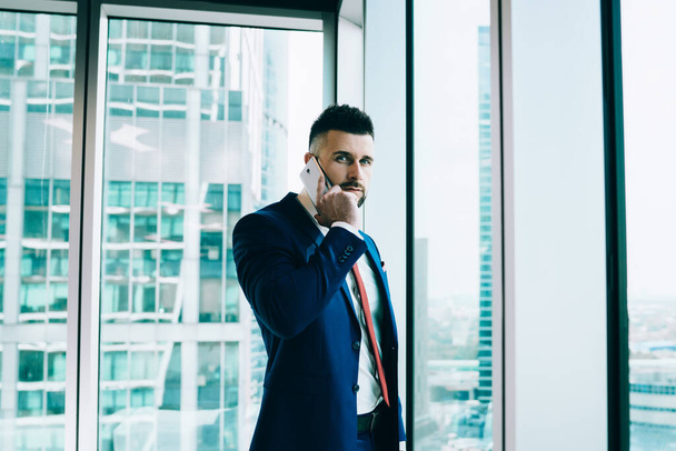 Confident focused professional entrepreneur in formal suit speaking on smartphone while standing near office window in cabinet on cityscape looking at camera - 写真・画像
