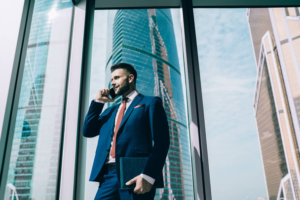 From below smiling confident man in elegant clothes with papers rejoicing in success while having conversation on phone with business partners in contemporary office - Foto, Imagen
