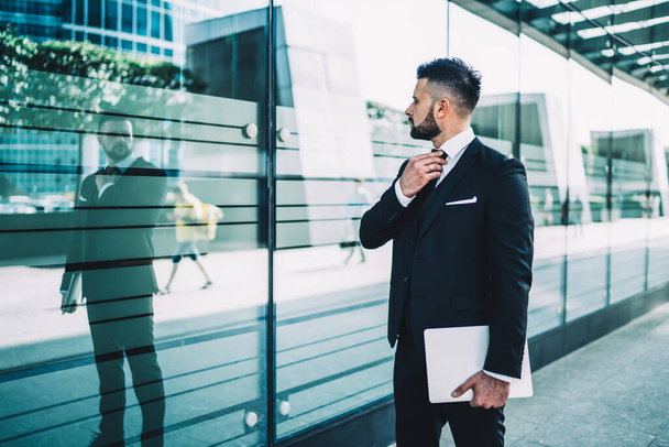 Confident businessman with tablet adjusting tie and looking at reflection in glass wall while standing outside modern building in downtown - Φωτογραφία, εικόνα