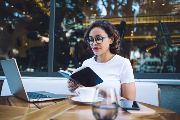 Serious young woman in casual outfit and glasses checking schedule in black organizer while working remotely in terrace of coffee shop - Fotó, kép