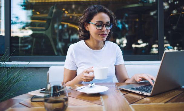 Smiling young woman wearing white t shirt and glasses and enjoying morning tea while typing on laptop in creative cafeteria - Fotografie, Obrázek