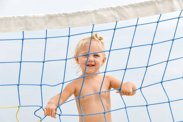 Niño alegre colgado en una red de voleibol contra el fondo del cielo. - Foto, imagen