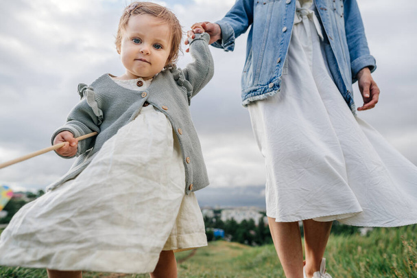 Imádnivaló kisbaba alkalmi ruhákat visel, kint sétál. Egy 1 éves kislány fogja az anyja kezét, és az első lépéseit a kamerába teszi.. - Fotó, kép