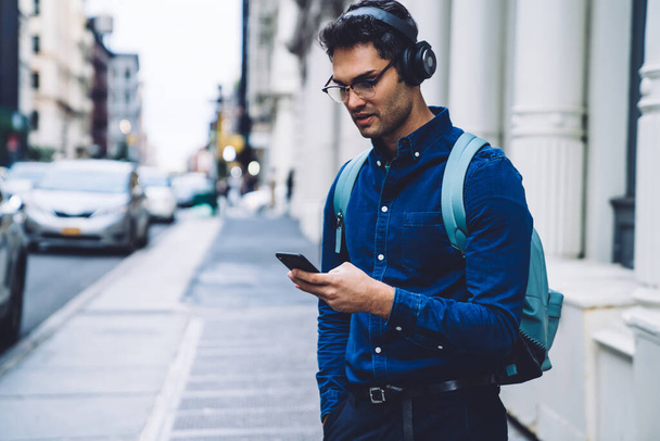 Confident handsome ethnic casual adult man in earphones and eyeglasses with backpack using smartphone while standing on urban street in daylight - Zdjęcie, obraz