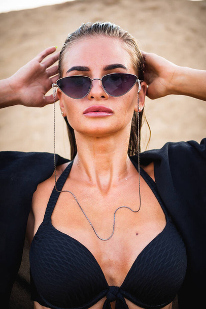 a beautiful brunette model in a black swimsuit and dark glasses, posing on the sand in front of the camera. Close-up and full-length shooting. - Foto, immagini