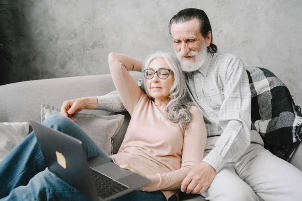 Senior couple sitting on couch with laptop - Фото, изображение