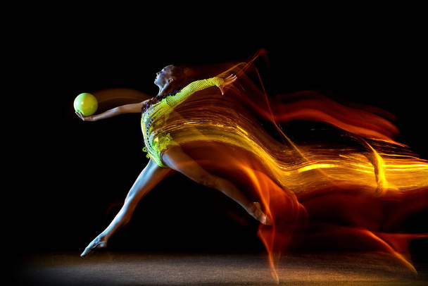 Portrait of young girl, rhythmic gymnastics artist in action isolated on dark studio background with mixed light. Concept of sport, action, aspiration, beauty - Fotó, kép
