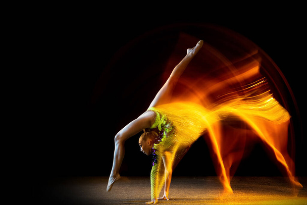 Portrait of young girl, rhythmic gymnastics artist in action isolated on dark studio background with mixed light. Concept of sport, action, aspiration, beauty - Foto, Imagem