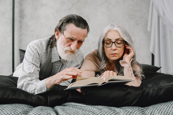 Senior couple reading book lying together on bed - Фото, изображение
