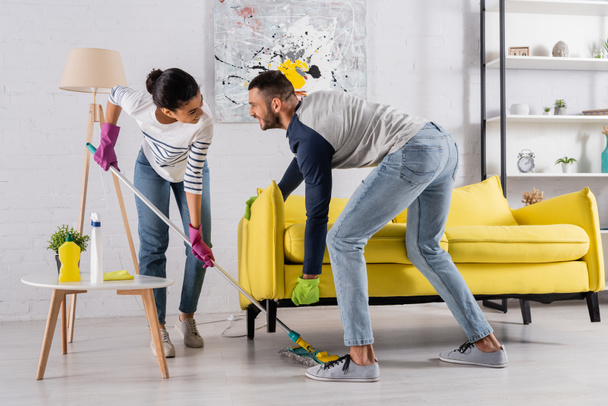 Allegro multietnico coppia pulizia pavimento sotto il divano a casa  - Foto, immagini