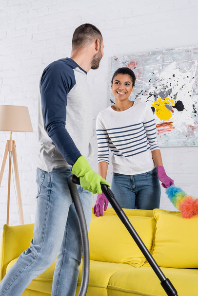Smiling african american woman with dust brush looking at boyfriend with vacuum cleaner  - Photo, Image