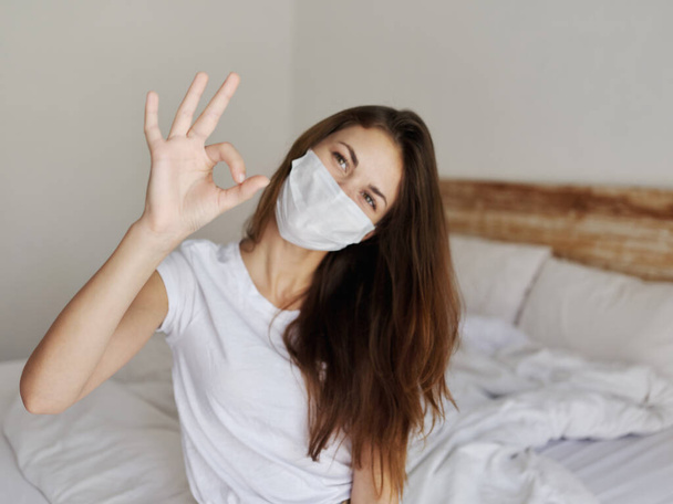 vrouw zittend op bed met medisch masker positieve handgebaren - Foto, afbeelding