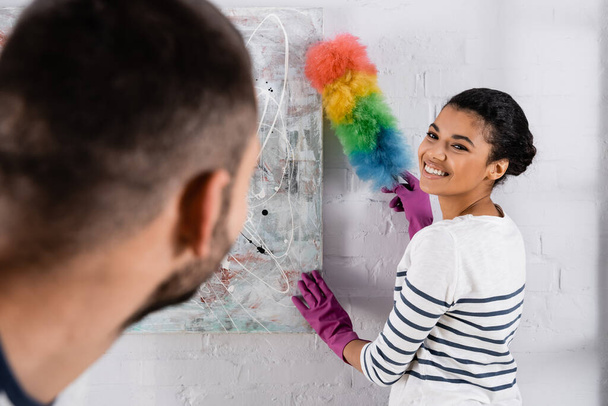 Smiling african american woman cleaning drawing with dust brush near blurred boyfriend  - Foto, immagini