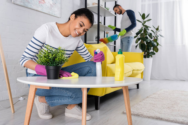 Positive african american woman cleaning coffee table near blurred boyfriend  - Photo, Image