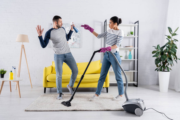 Side view of interracial couple with detergent and vacuum cleaner having fun in living room  - Fotoğraf, Görsel