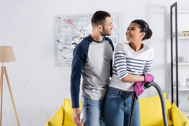 Smiling interracial couple with vacuum cleaner and detergent at home  - Fotografie, Obrázek