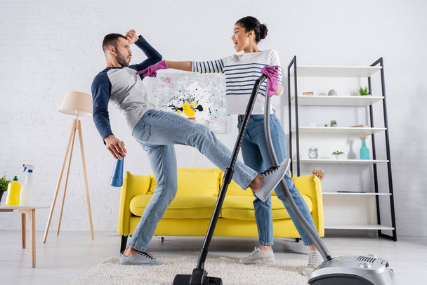 Side view of african american woman with vacuum cleaner pointing at boyfriend with detergent  - Fotó, kép