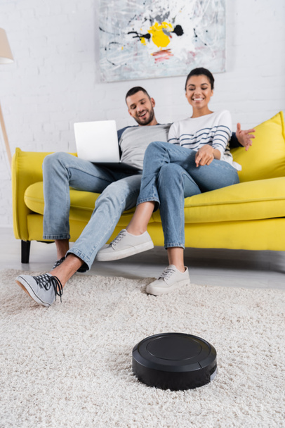 Robotic vacuum cleaner near blurred interracial couple with laptop  - Photo, Image