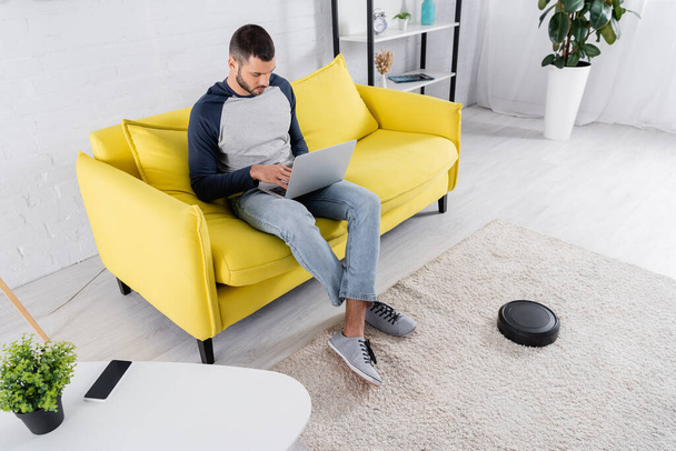 Young man using laptop near robotic vacuum cleaner at home  - Photo, Image