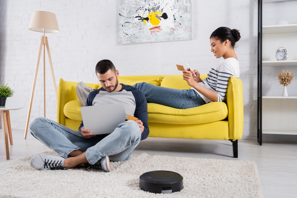 Smiling multiethnic couple using devices and credit cards near robotic vacuum cleaner  - Photo, Image