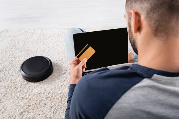 Back view of blurred man holding credit card and laptop near robotic vacuum cleaner  - Photo, Image
