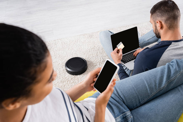Robotic vacuum cleaner on carpet near interracial couple using devices and credit card  - Photo, Image