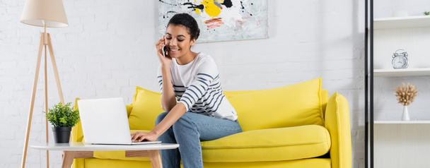Lächelnde Afroamerikanerin im Gespräch mit Smartphone und Laptop im Wohnzimmer, Banner  - Foto, Bild