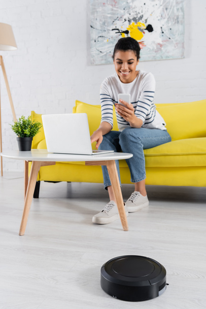 Robotic vacuum cleaner near blurred african american freelancer with smartphone and laptop  - Foto, imagen