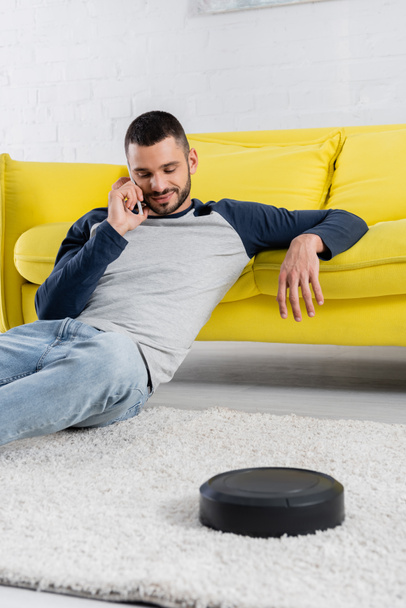 Young man talking on smartphone near blurred robotic vacuum cleaner  - Foto, immagini