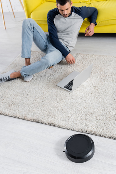 High angle view of robotic vacuum cleaner on floor and freelancer using laptop on background  - Foto, afbeelding