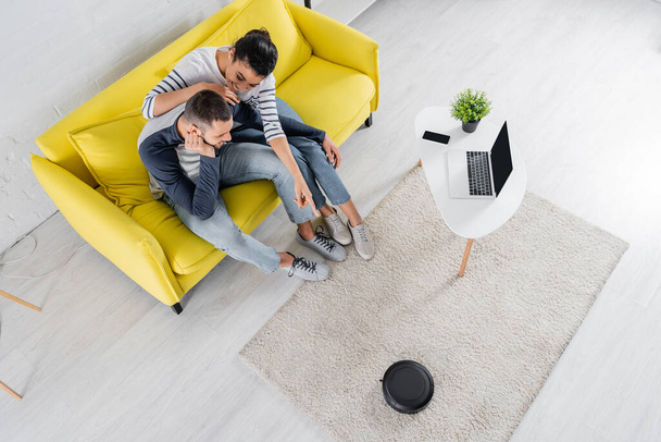 Overhead view of smiling african american woman pointing at robotic vacuum cleaner near boyfriend  - Zdjęcie, obraz