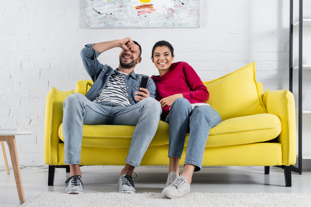 Positive multiethnic couple with remote controller sitting on couch  - Foto, immagini