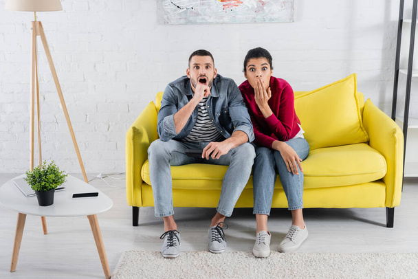Amazed interracial couple with remote controller looking at camera near devices on coffee table  - Photo, image