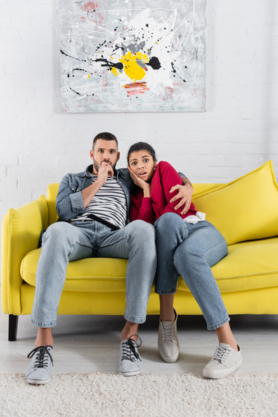 Scared interracial couple looking at camera near remote controller on couch  - Foto, afbeelding