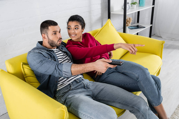 Smiling african american woman pointing with finger near amazed boyfriend with remote controller  - 写真・画像