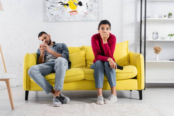 Mujer afroamericana asustada sosteniendo controlador remoto cerca de novio usando teléfono inteligente en el sofá  - Foto, imagen