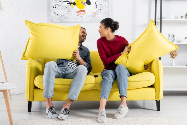 Cheerful multiethnic couple holding pillows on couch  - Valokuva, kuva