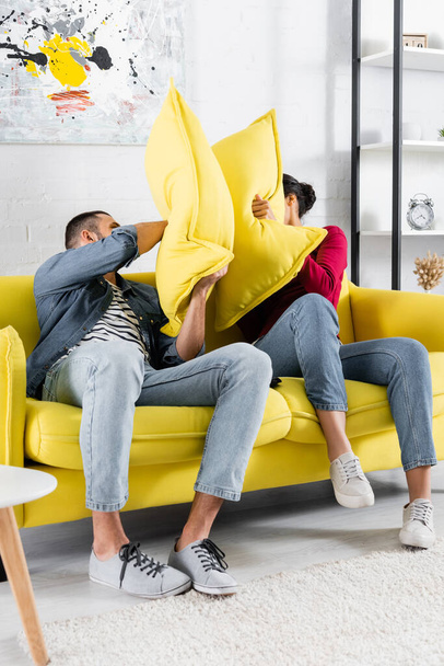 Multiethnic couple pillow fighting on couch in living room  - Φωτογραφία, εικόνα