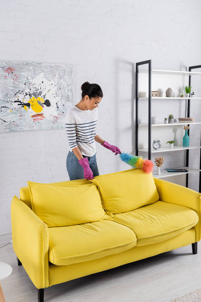 Seitenansicht einer lächelnden Afroamerikanerin in Gummihandschuhen mit Staubbürste beim Putzen der Couch  - Foto, Bild