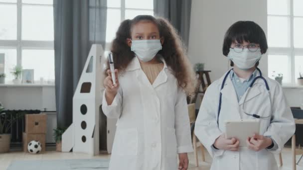 Porträt mit Handheld-Ortung von 8-jährigem Mädchen und 6-jährigem Jungen in Arztkostümen und Gesichtsmasken mit Tablette und Spritze, die mit Medikamenten gefüllt sind und zu Hause in die Kamera schauen - Filmmaterial, Video