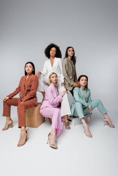 full length of interracial models in stylish pastel suits sitting while posing on grey - Foto, Imagem