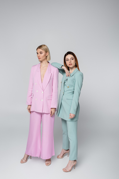 full length of blonde and redhead women in pink and blue pastel suits posing on grey - Фото, зображення